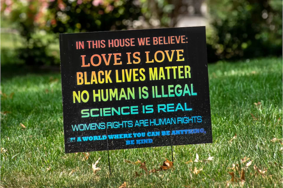 A rainbow colored sign planted in the front yard of a typical suburban home reads, "In this house we believe love is love, black lives matter, no human is illegal, science is real, women's rights are human rights. In a world where you can be anything, be kind."